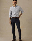 A man modeling a light blue Faherty long-sleeve button-down shirt, dark navy trousers, a brown belt, and brown leather loafers, standing against a textured beige studio backdrop.