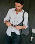 A man wearing a Faherty white and blue button-down shirt and dark blue jeans stands against a dark wood-paneled wall, holding a pair of sunglasses in his hands while carrying a gray shoulder bag.