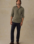 A man modeling a long-sleeve button-up shirt in olive green with dark navy pants and brown shoes, standing against a textured beige studio backdrop.