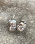 A pair of polished sterling silver huggie earrings with a heart-shaped clear cubic zarconia gemstones, displayed on a textured gray and white crystalline surface.