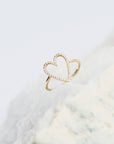 A solid 14k gold heart-shaped halo ring with small diamond gemstones along the outline, displayed on a textured white surface against a soft white background.