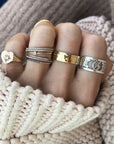 A hand showcasing many different rings in silver and gold vermeil. With a hammered textured cigar band on the middle finger, with a sweater texture in the background.