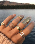 A hand wearing multiple JNB Style rings, with a Stella Ring, a hammered cigar band in gold vermeil, on the middle finger. With a lake and mountains in the background.