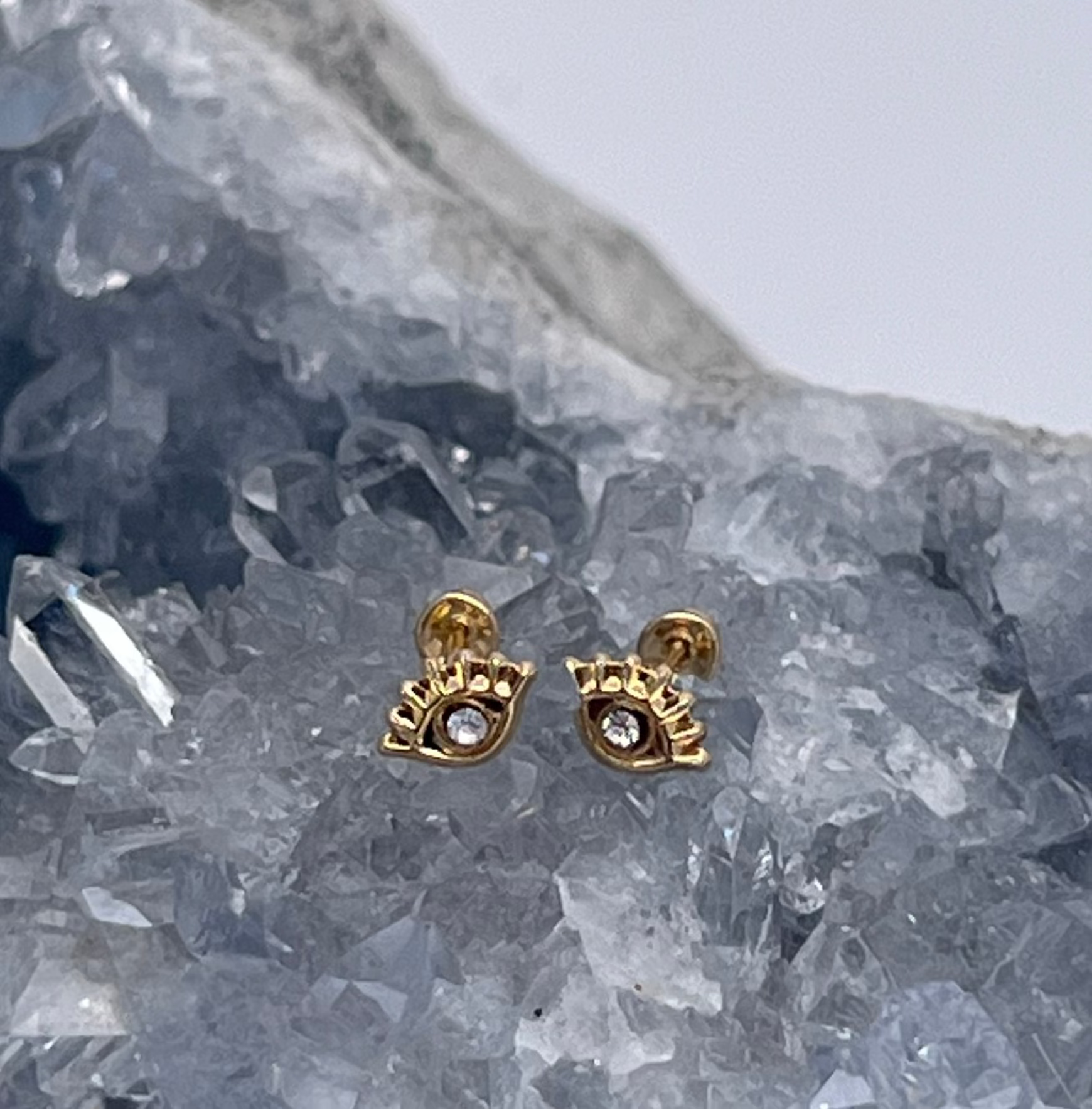 A pair of 114k yellow gold evil eye stud earrings with a diamond center, displayed on a crystal.