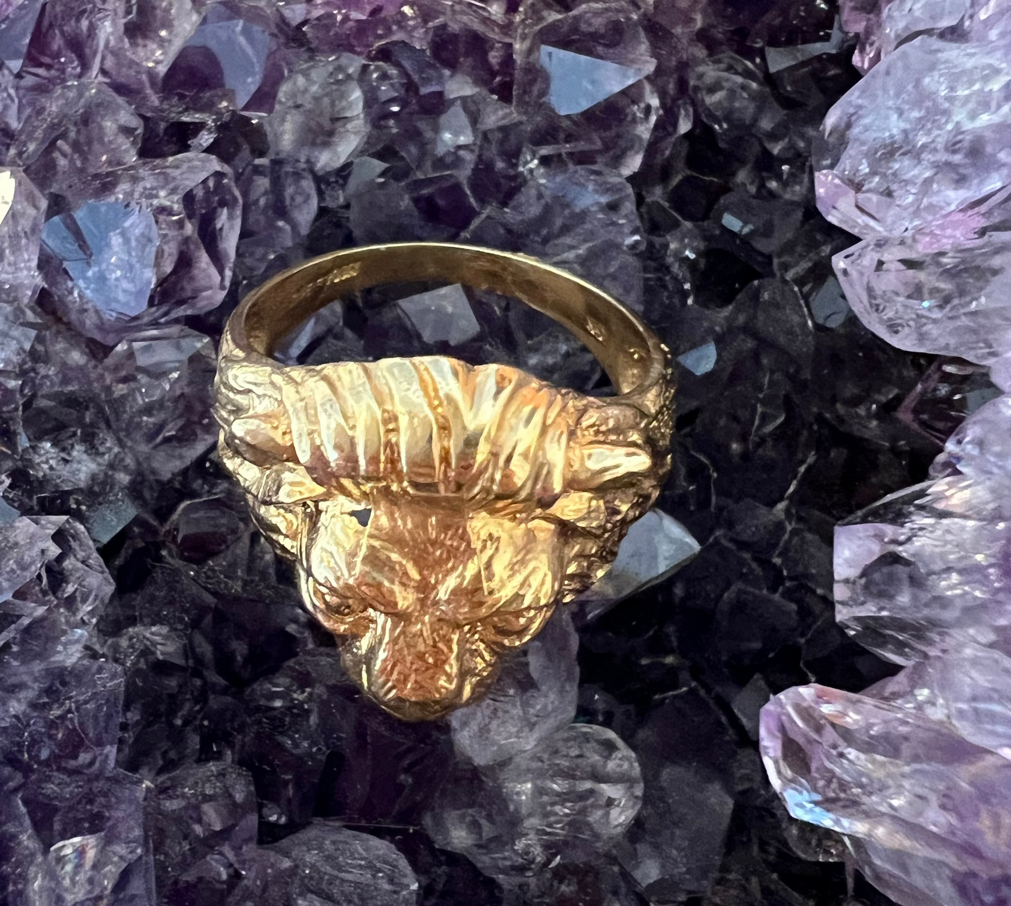 The top view of the Aslan King of Beasts Gold Vermeil Lion Ring. Showing the thick band, and detailing with the lions furrowed brow and mane. Against an Amethyst crystal background. 