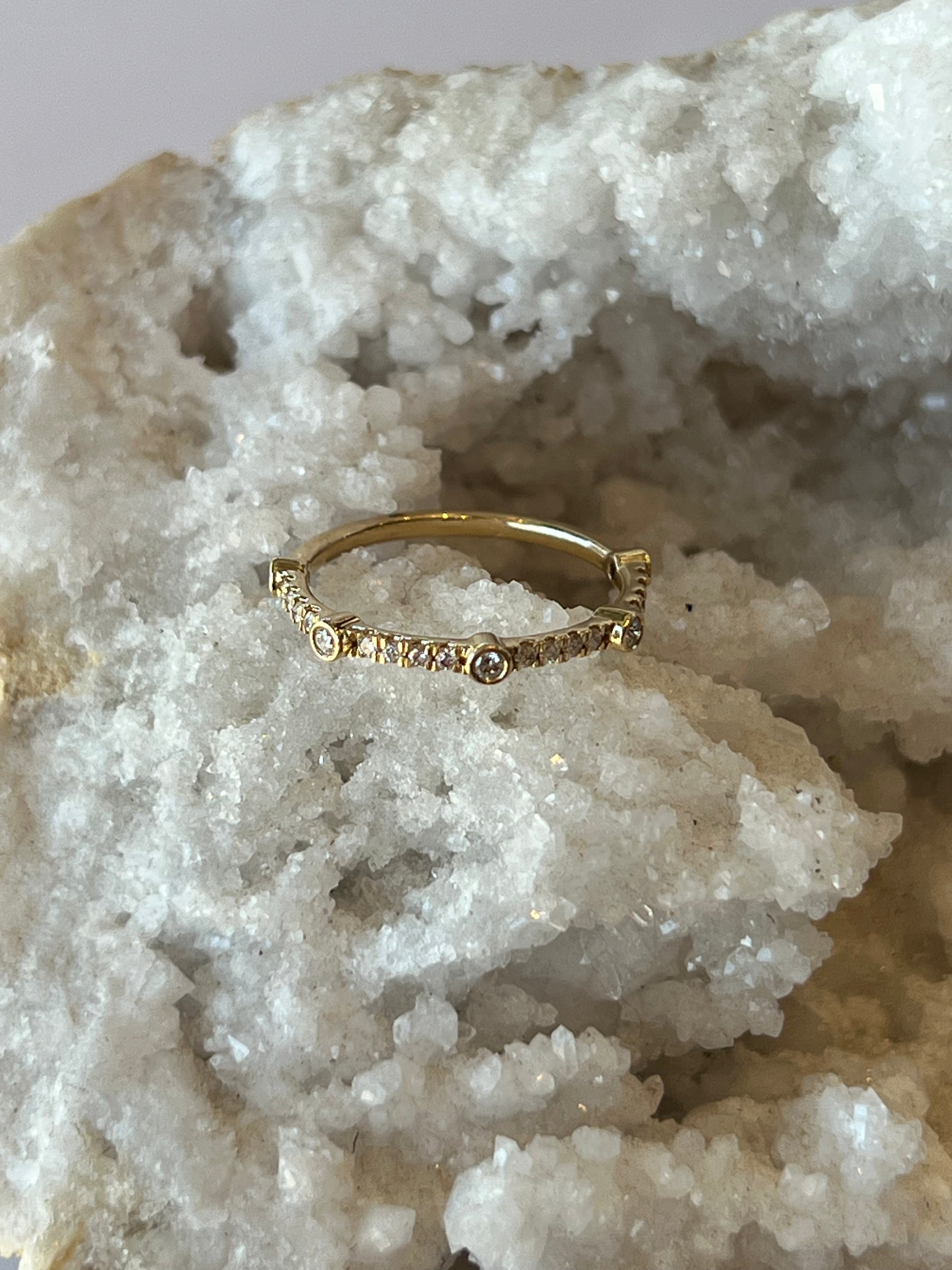 A solid 14k gold band with multiple brilliant cut diamonds set in bezel settings, with pave diamonds in between, creating a unique diamond band. Displayed on a bed of white crystals.