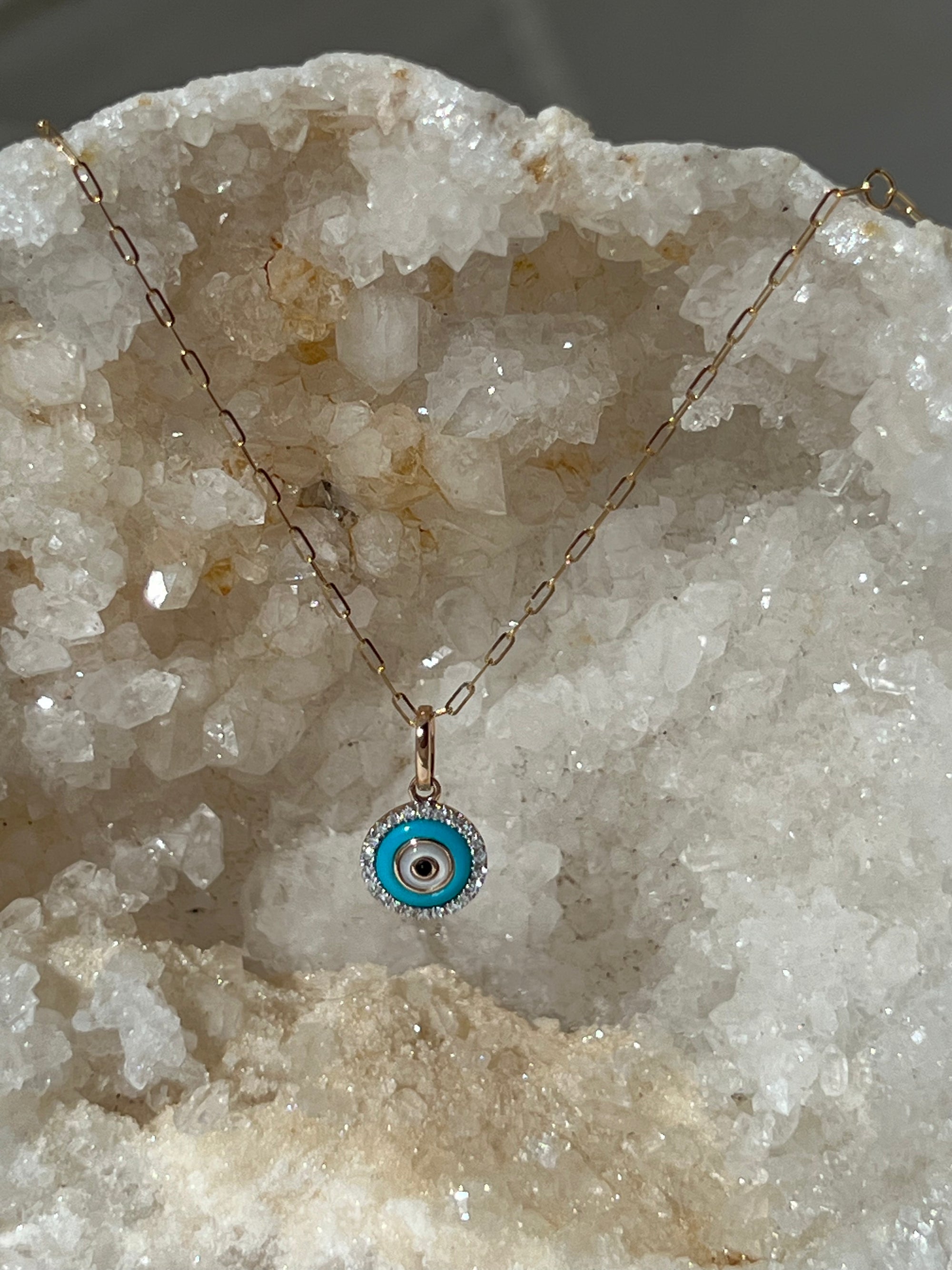 A 14k gold box chain necklace with an evil eye pendant made of gold, turquoise, diamonds, onyx to offer protection and spiritual meaning displayed on a crystal surface.