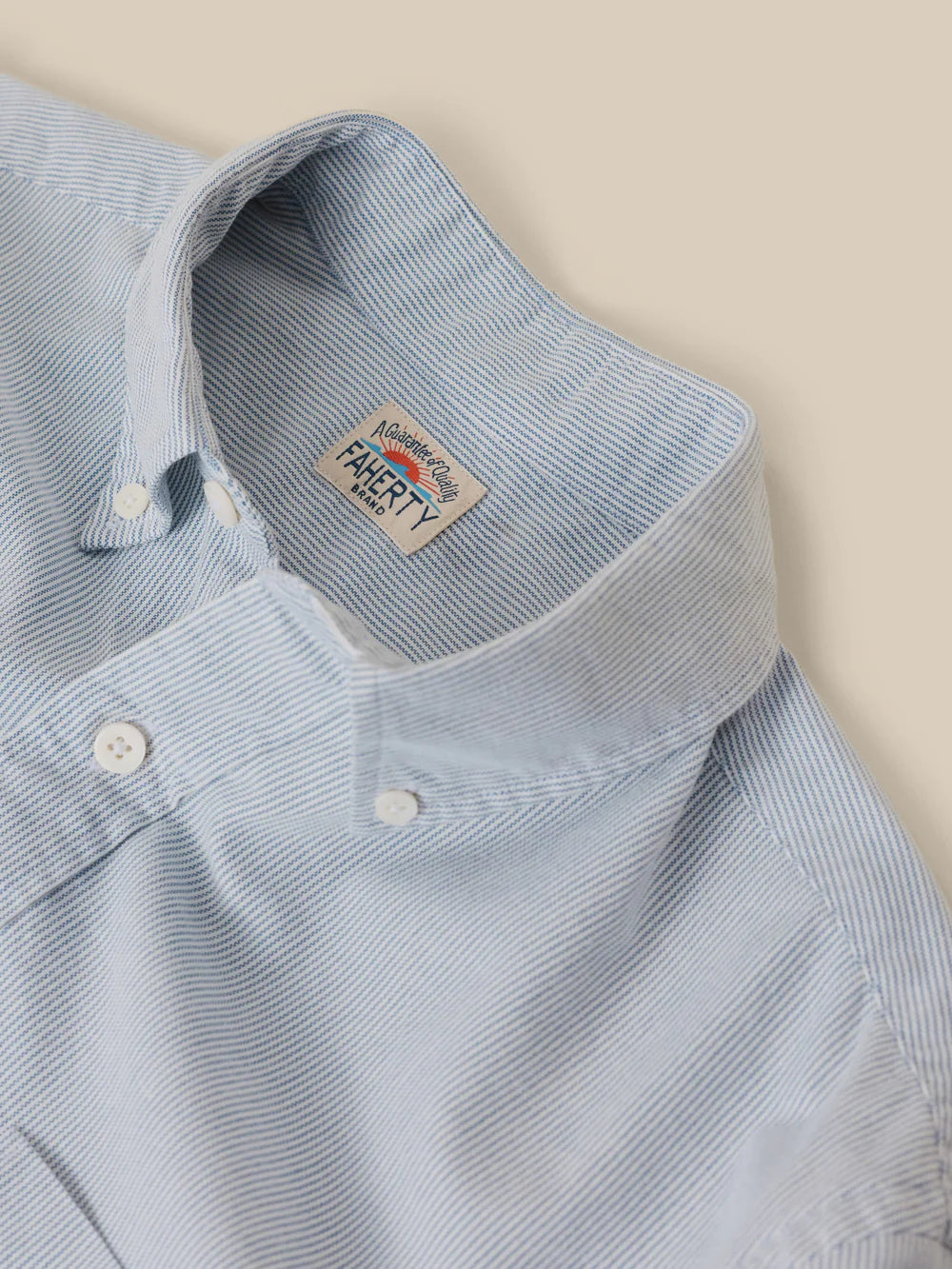 Close-up of a light blue and white micro-striped button-down shirt collar, showing the 'FAHERTY' brand label inside the neck, set against a solid beige background.