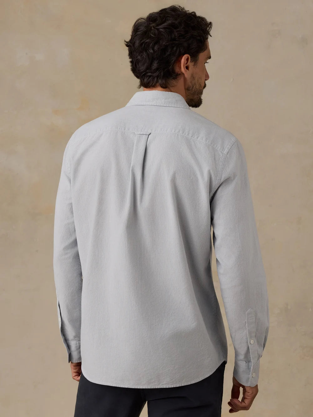 A man viewed from the back wearing a light blue striped Faherty Oxford long-sleeve button-down shirt and dark trousers, standing against a textured beige wall.