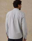 A man viewed from the back wearing a light blue striped Faherty Oxford long-sleeve button-down shirt and dark trousers, standing against a textured beige wall.