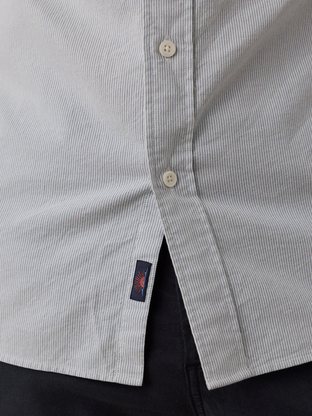 Close-up of a light blue and white striped organic cotton button-down shirt with a small navy brand tag featuring a red sun logo, worn over dark pants.