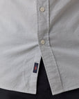 Close-up of a light blue and white striped organic cotton button-down shirt with a small navy brand tag featuring a red sun logo, worn over dark pants.