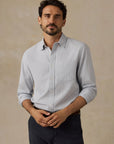A man wearing a Faherty light blue long-sleeve button-down shirt and dark trousers, standing against a textured beige background.