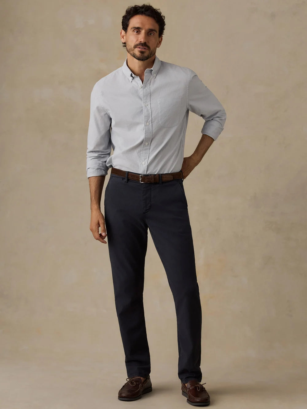 A man modeling a light blue Faherty long-sleeve button-down shirt, dark navy trousers, a brown belt, and brown leather loafers, standing against a textured beige studio backdrop.