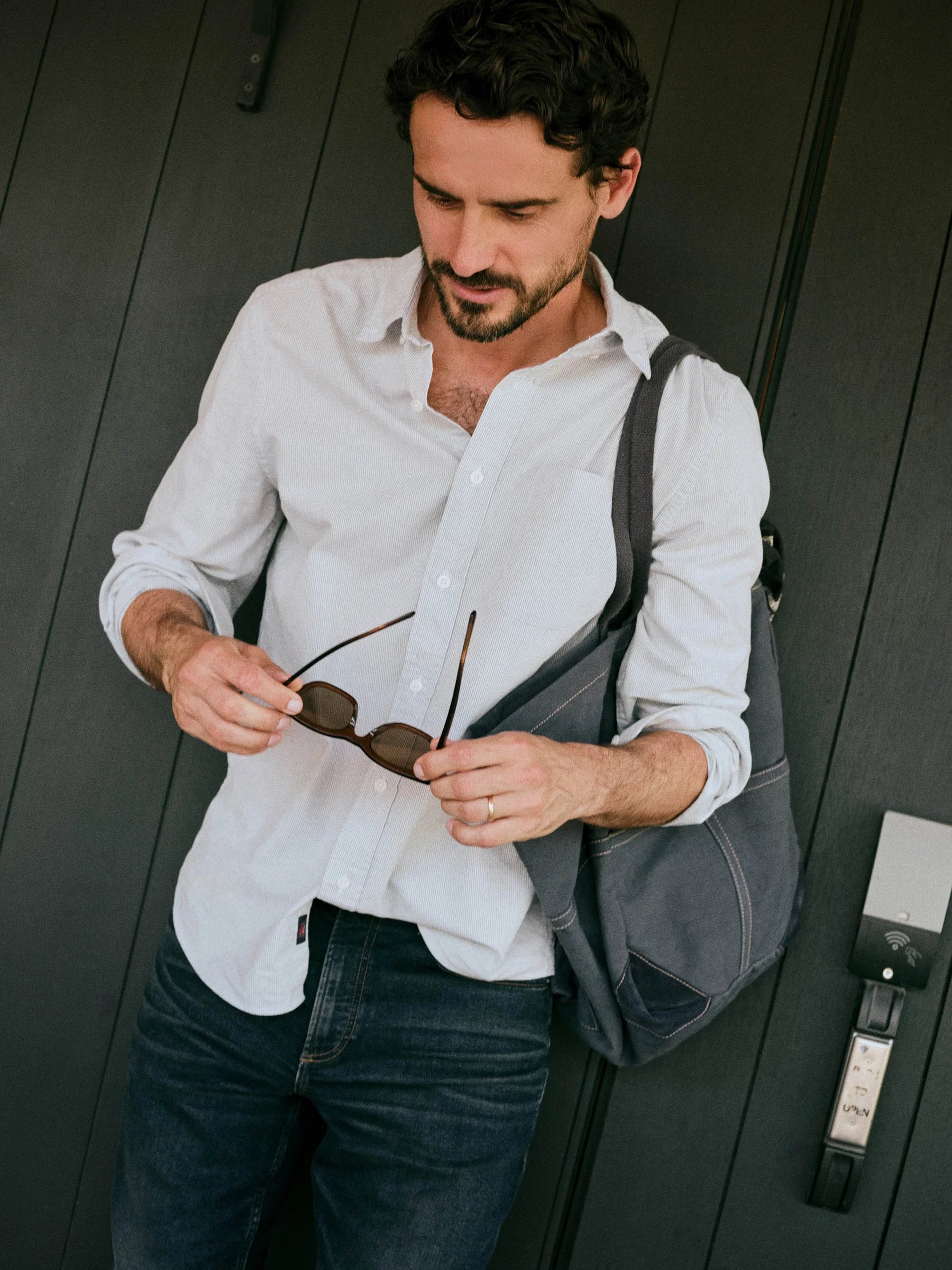 A man wearing a Faherty white and blue button-down shirt and dark blue jeans stands against a dark wood-paneled wall, holding a pair of sunglasses in his hands while carrying a gray shoulder bag.