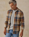 A man wearing a brown, beige, and blue plaid button-up shirt over a light gray t-shirt, paired with light blue jeans, standing against a textured beige studio backdrop.