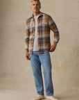A man wearing a brown, beige, and blue plaid button-up shirt over a light gray t-shirt, paired with light blue jeans and off-white sneakers, standing against a textured beige studio backdrop.