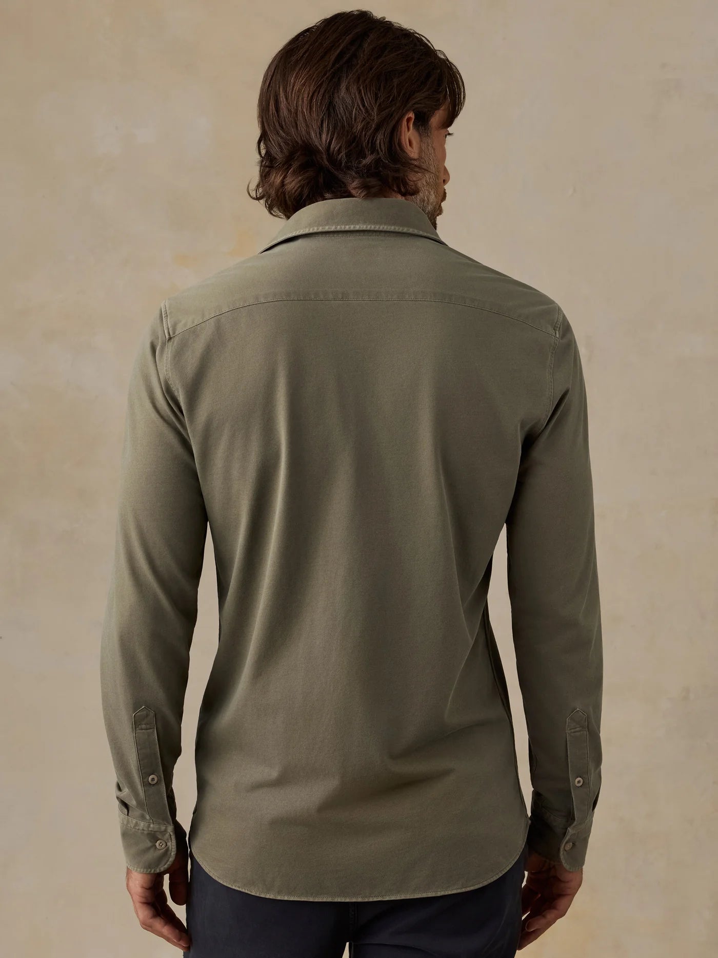 A man viewed from the back wearing a muted sage green long-sleeve button-down shirt, paired with dark navy pants, against a textured beige background.