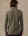 A man viewed from the back wearing a muted sage green long-sleeve button-down shirt, paired with dark navy pants, against a textured beige background.