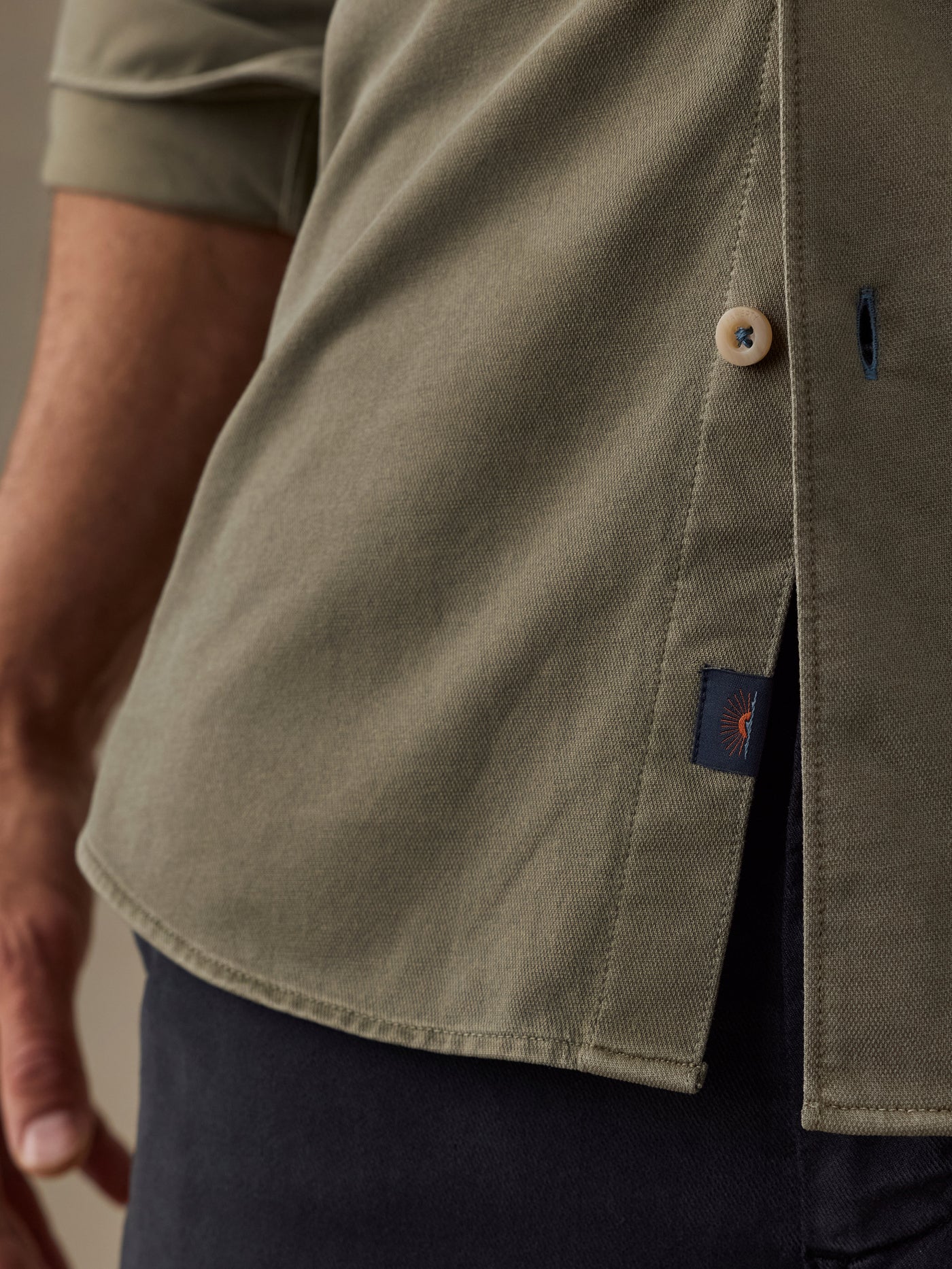 Close-up of a man wearing an olive green short-sleeve button-up shirt with a side slit and a small navy brand tab near the hem.