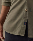 Close-up of a man wearing an olive green short-sleeve button-up shirt with a side slit and a small navy brand tab near the hem.