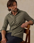 A man wearing an olive green long-sleeve button-up shirt and dark navy pants, sitting on a wooden chair against a textured beige wall.