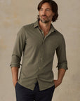 A man wearing a muted olive green long-sleeve button-down shirt with rolled-up sleeves and dark navy pants, standing against a textured beige studio backdrop.