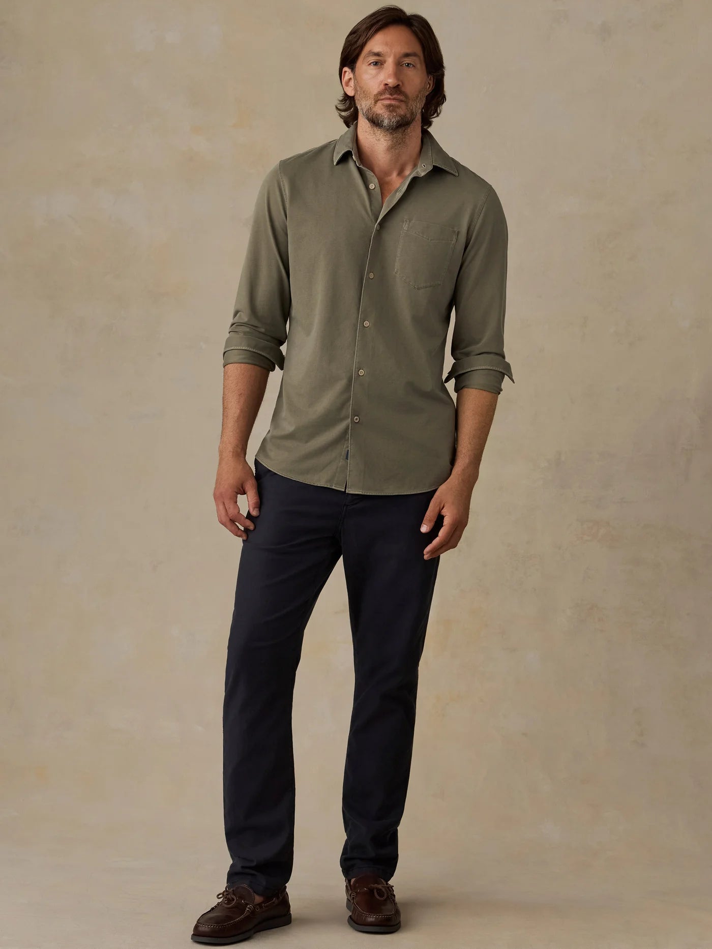 A man modeling a long-sleeve button-up shirt in olive green with dark navy pants and brown shoes, standing against a textured beige studio backdrop.