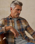 A man wearing a brown, beige, and blue plaid button-up shirt over a light gray t-shirt, paired with light blue jeans, sitting against a textured beige studio backdrop.