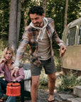 A man outdoors wearing a boulder creek plaid button down, setting down a red kettle, wth a woman in a pink Faherty button down behind him. 
