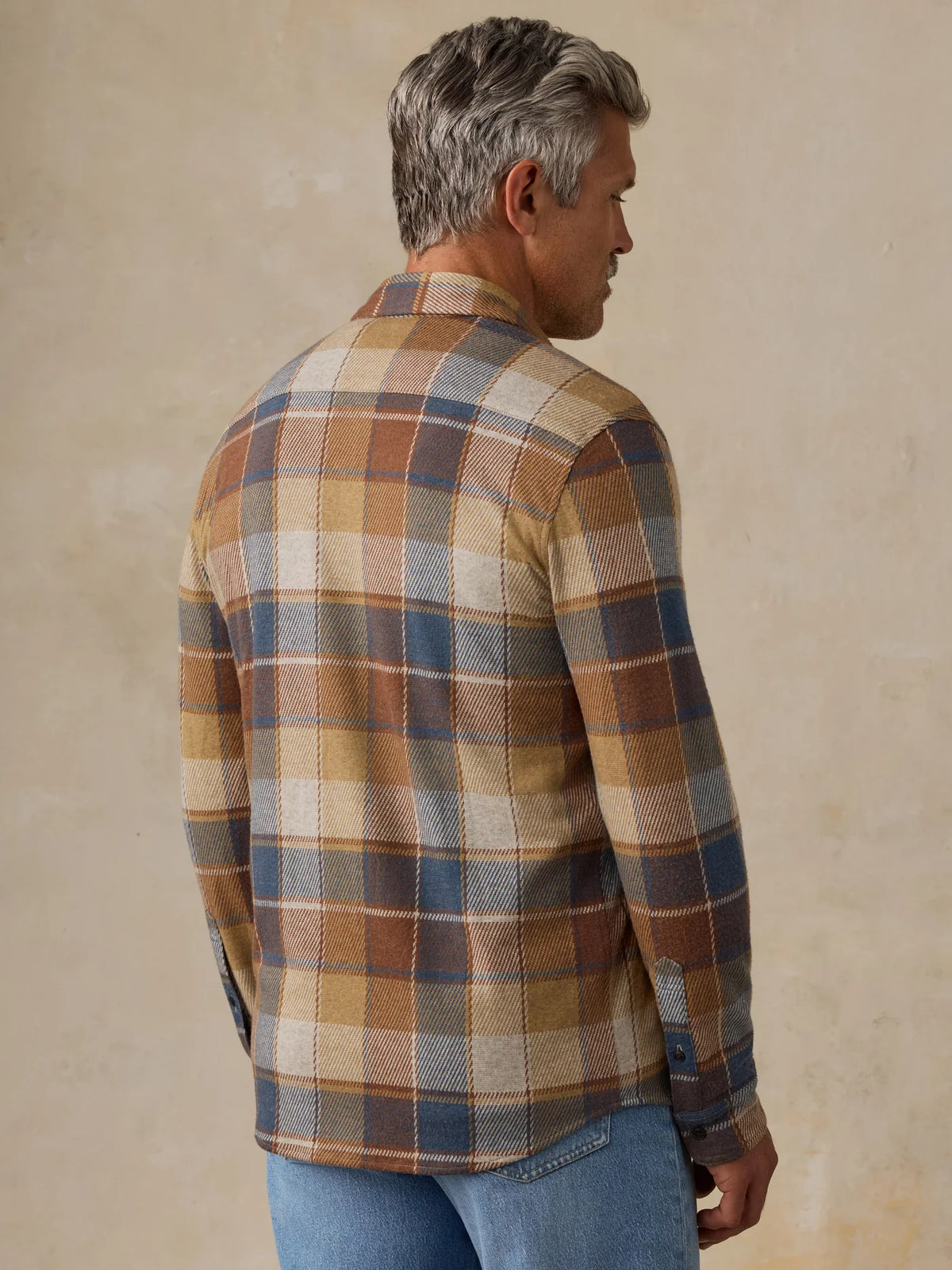 A man wearing a brown, beige, and blue plaid button-up shirt viewed from the back, paired with light blue jeans and off-white sneakers, standing against a textured beige studio backdrop.