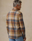 A man wearing a brown, beige, and blue plaid button-up shirt viewed from the back, paired with light blue jeans and off-white sneakers, standing against a textured beige studio backdrop.