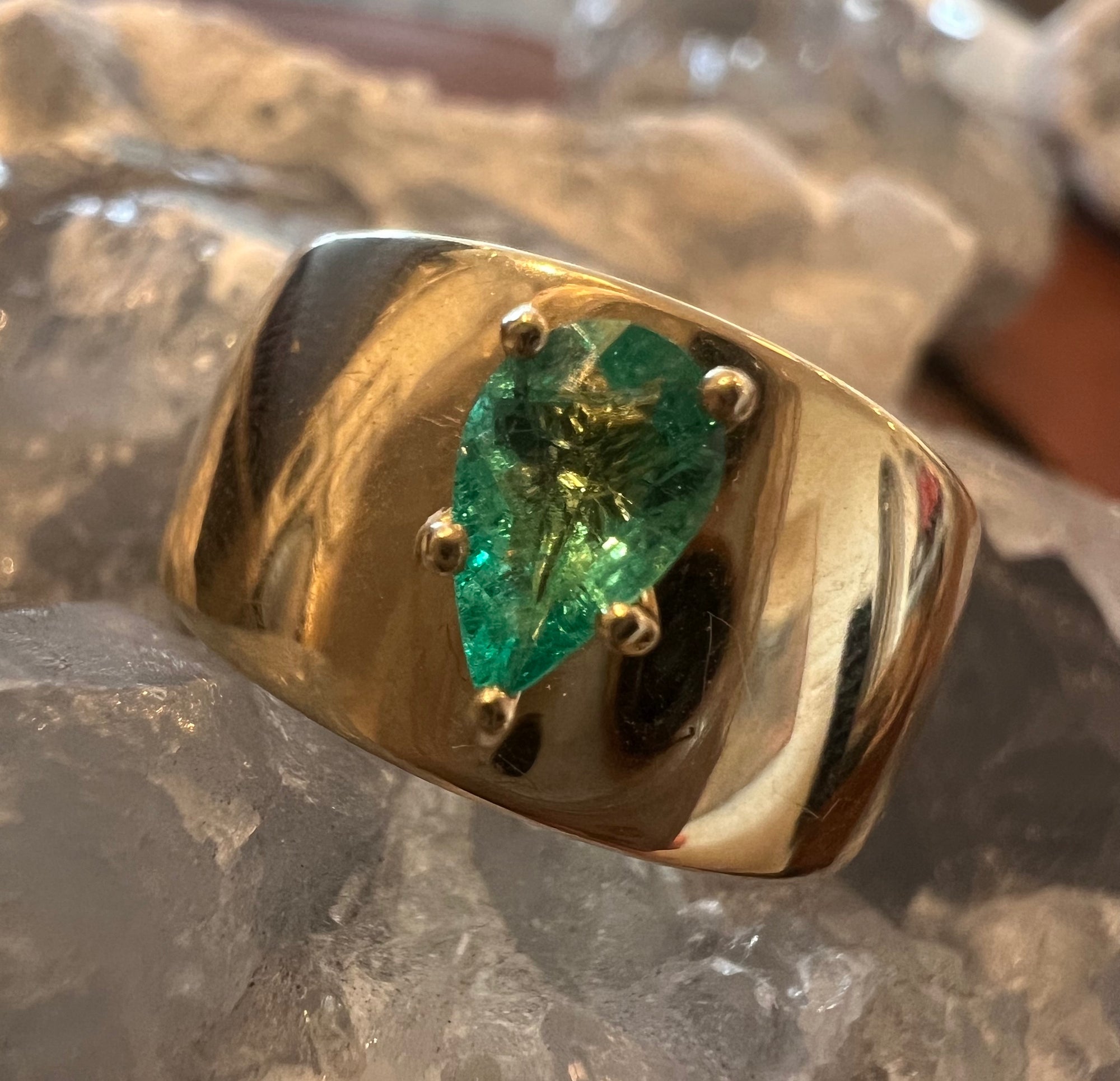 A wide 14k yellow gold ring featuring a pear-cut GIA Certified green emerald gemstone in a four-prong setting, displayed on a bed of clear quartz crystals.
