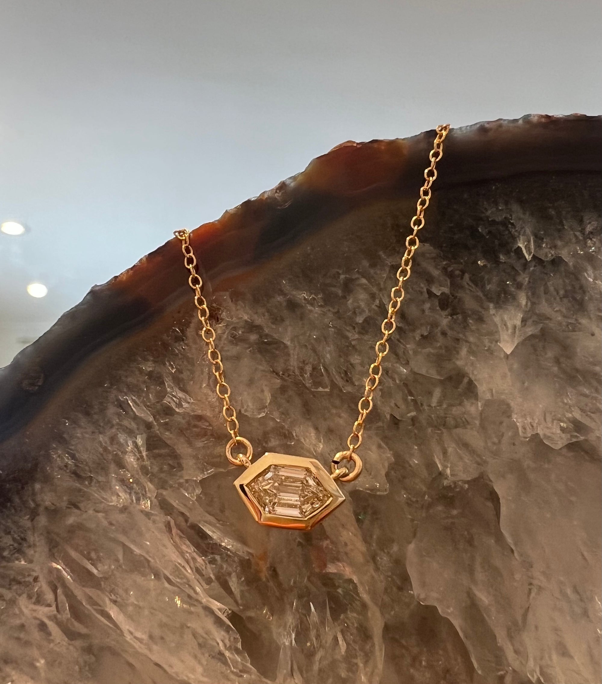 14k yellow hold necklace with a bezel set hexagon diamond. The diamond pendant is set securely at the center of a cable chain. The background is a quartz crystal.