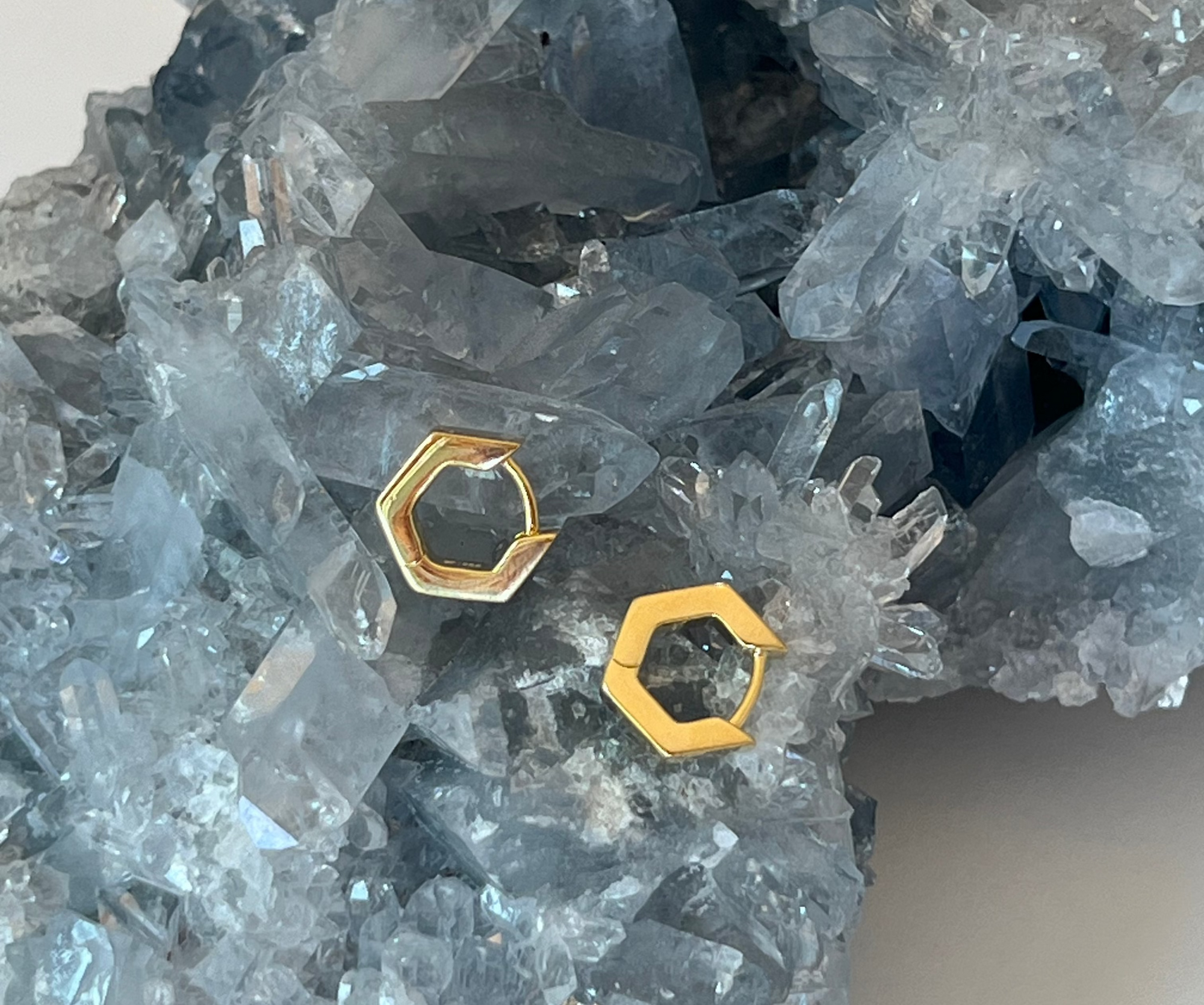 Two small gold geometric hexagon huggie earrings made in California on a blue-gray crystal cluster against a light neutral background.