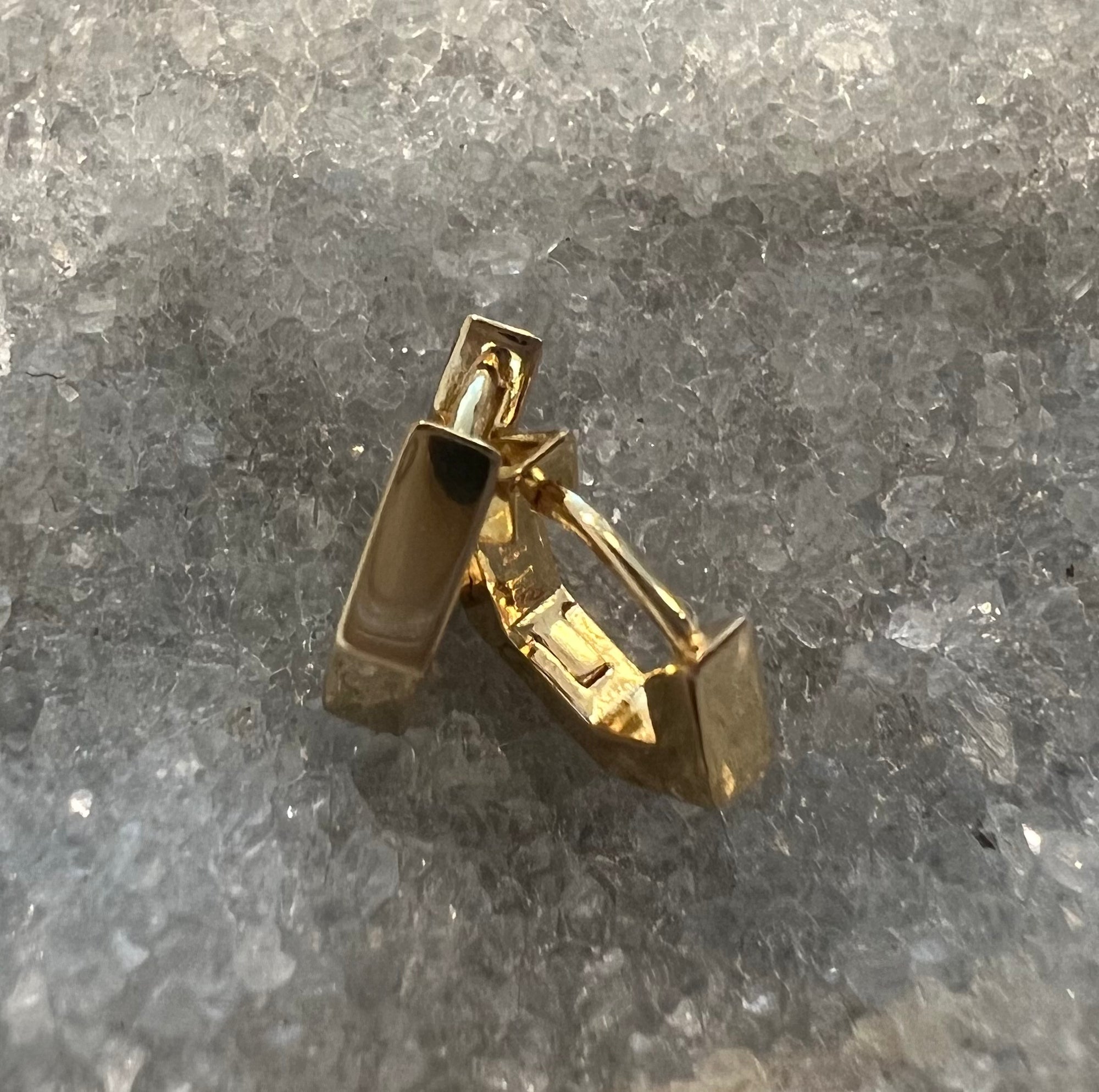 A pair of gold vermeil hexagon huggie hoop earrings designed by Jacqueline Nicole Walton with rectangular barbell-like details, displayed on a textured gray crystal surface.