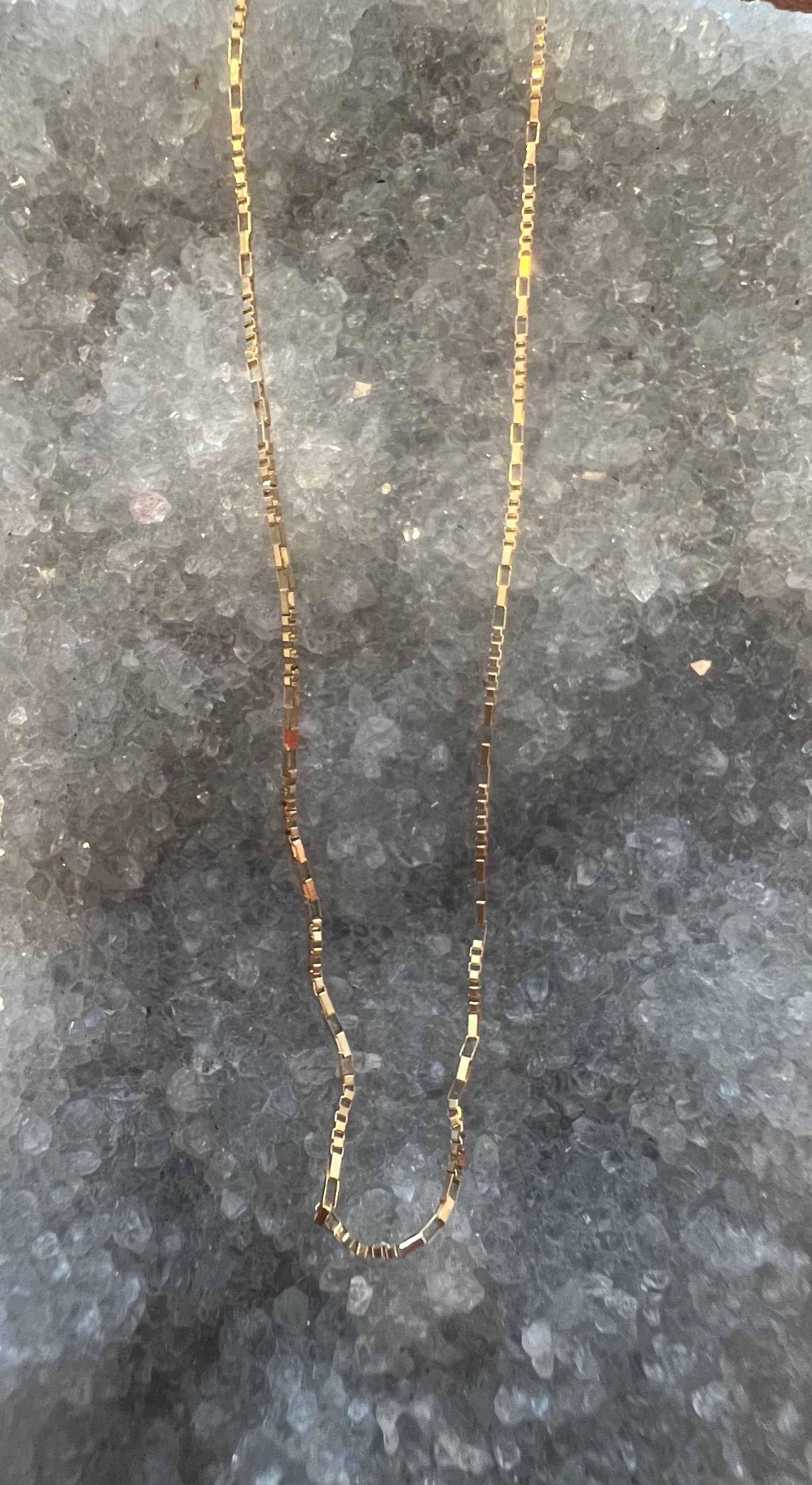 A delicate and unique solid 14k gold box chain necklace laid flat on a textured gray stone surface.