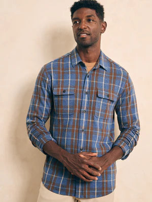 A man wearing a long-sleeved Faherty Legends Sweater Shirt in Alpine Lake Plaid shirt in blue and brown colors with a buttoned front and two chest pockets.
