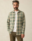 A male model wearing a Faherty  Forest Drive Plaid long-sleeved plaid flannel shirt in shades of green and beige with a white t-shirt underneath. 