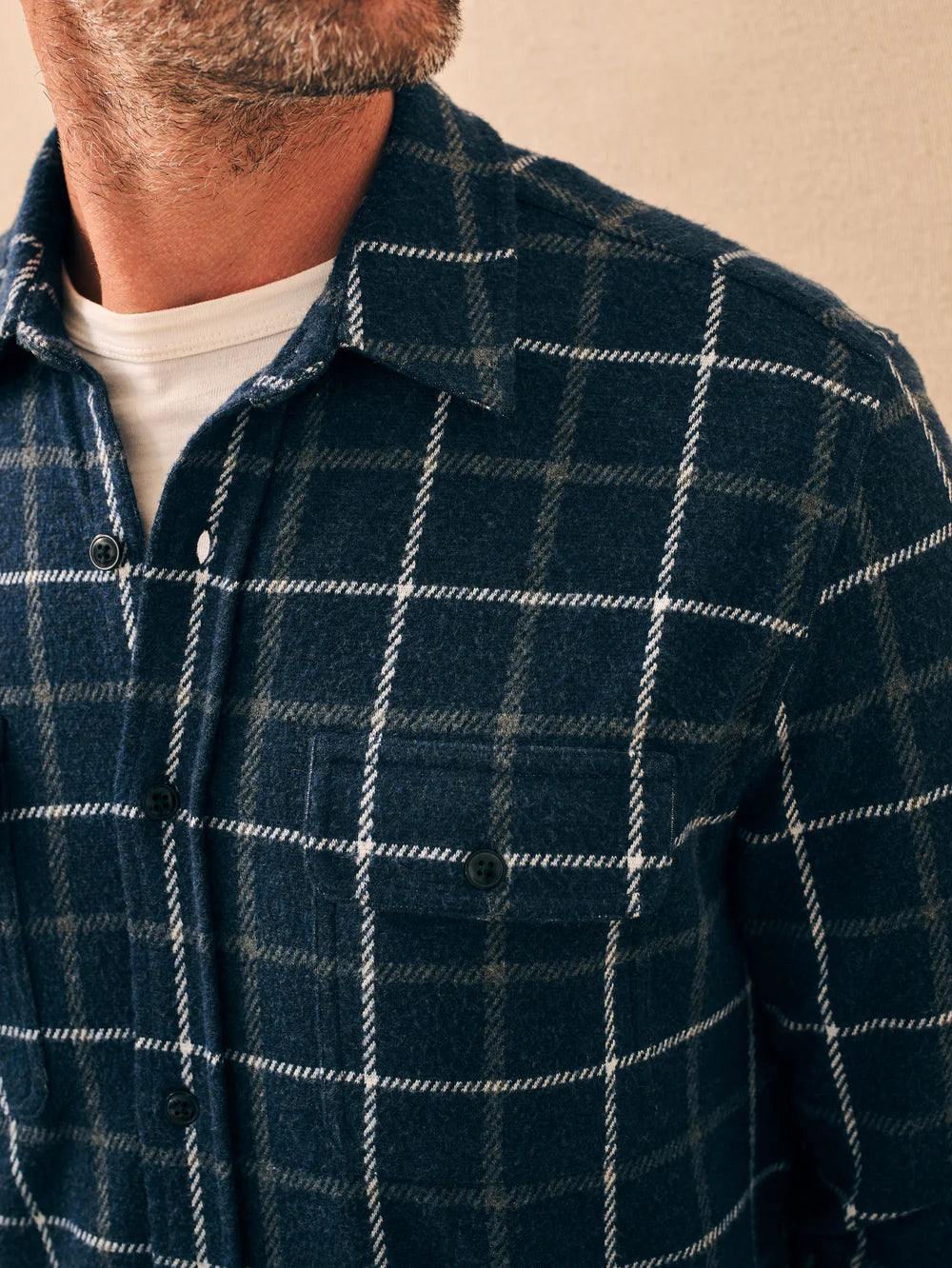 A man wearing a navy, grey, and white plaid button down shirt, with a a detail pocket and buttons. The shirt is by Faherty, and it is their Legend Sweater Shirt in Low Rider Blue.