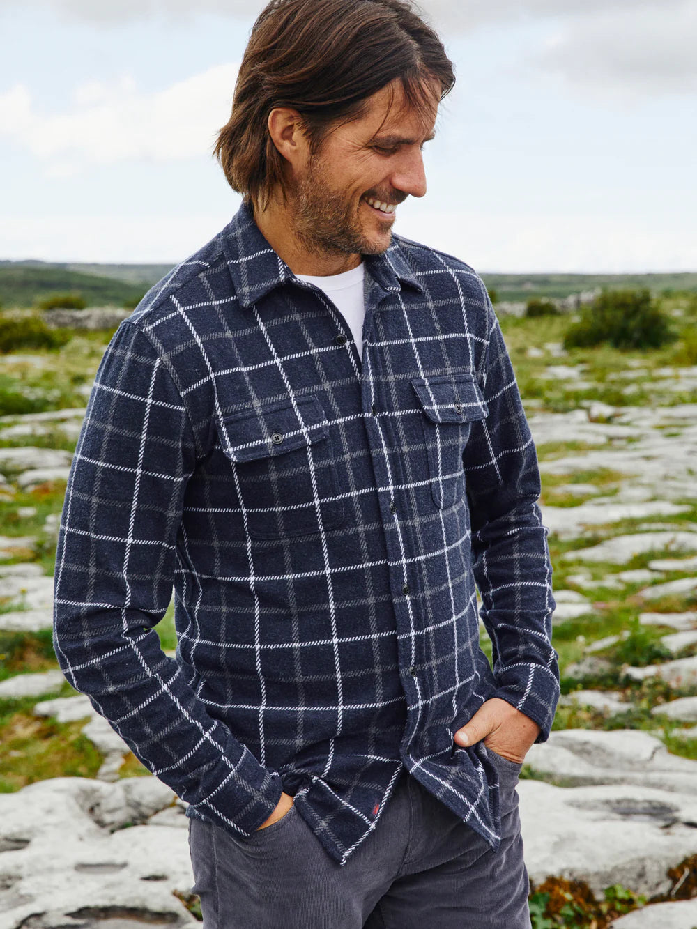 A man wearing a Faherty Legend Sweater Shirt in Low Rider Blue long-sleeved blue flannel shirt with a plaid navy,grey, and white buttoned collar, and front pockets.