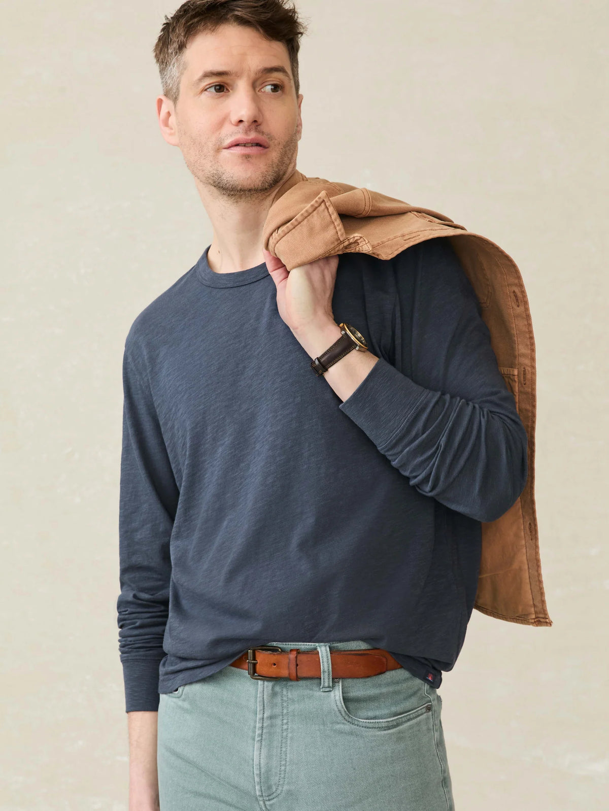 A man wearing a Faherty Long-Sleeve Sunwashed Tee in Dune Navy and light jeans, holding a brown jacket against a beige background