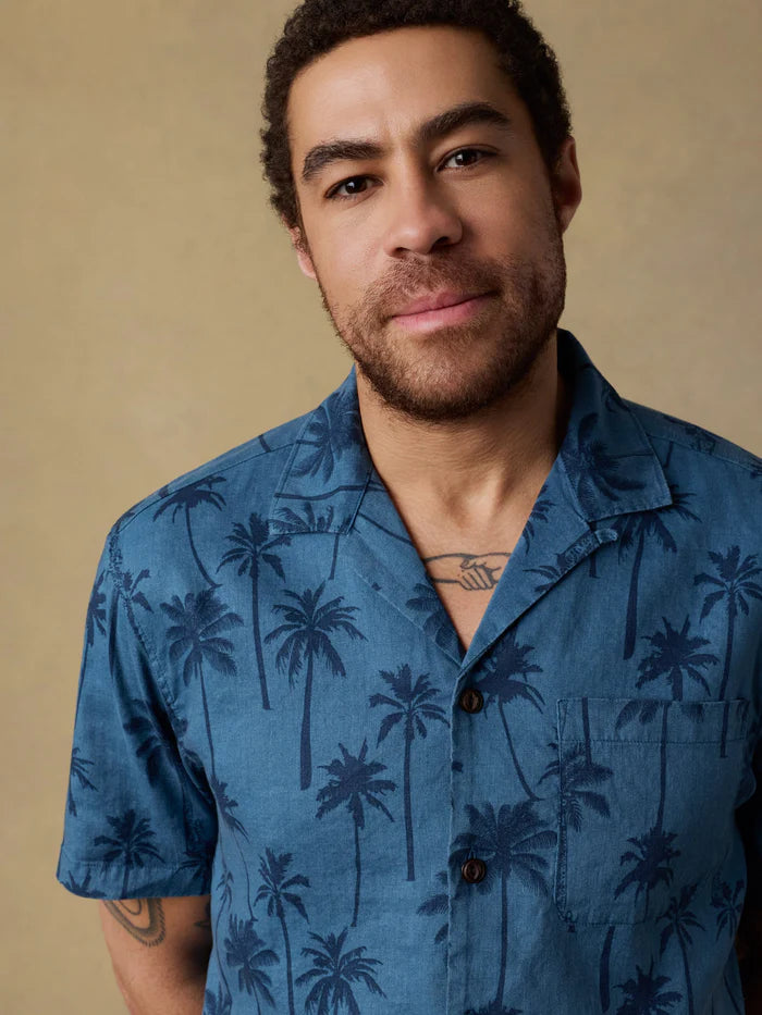 A man wearing a Faherty blue shirt with palm tree pattern against a beige background. 