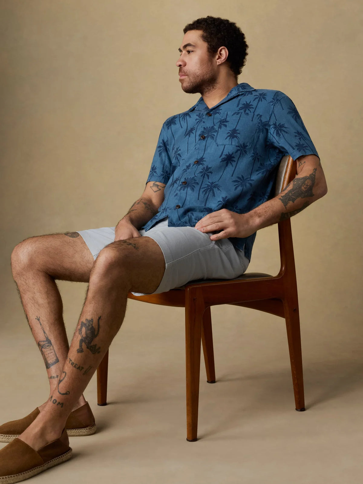 A man sitting on a chair wearing a blue patterned shirt with palm trees and light gray shorts against a beige background. 