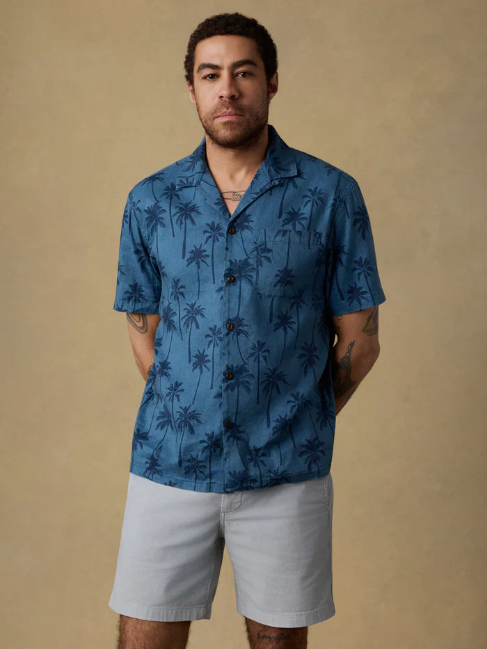 A man wearing a blue palm-tree patterned shirt and light gray shorts against a beige background.