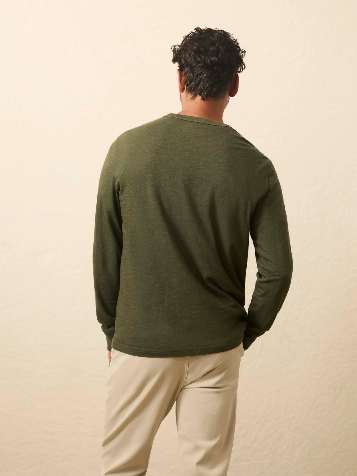 The back of a person wearing a green long-sleeve shirt and beige pants against a beige background