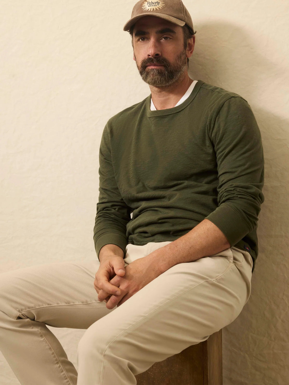 A man wearing a green Faherty Long-Sleeve Sunwashed Tee in Fall Evergreen shirt and beige pants against a beige background