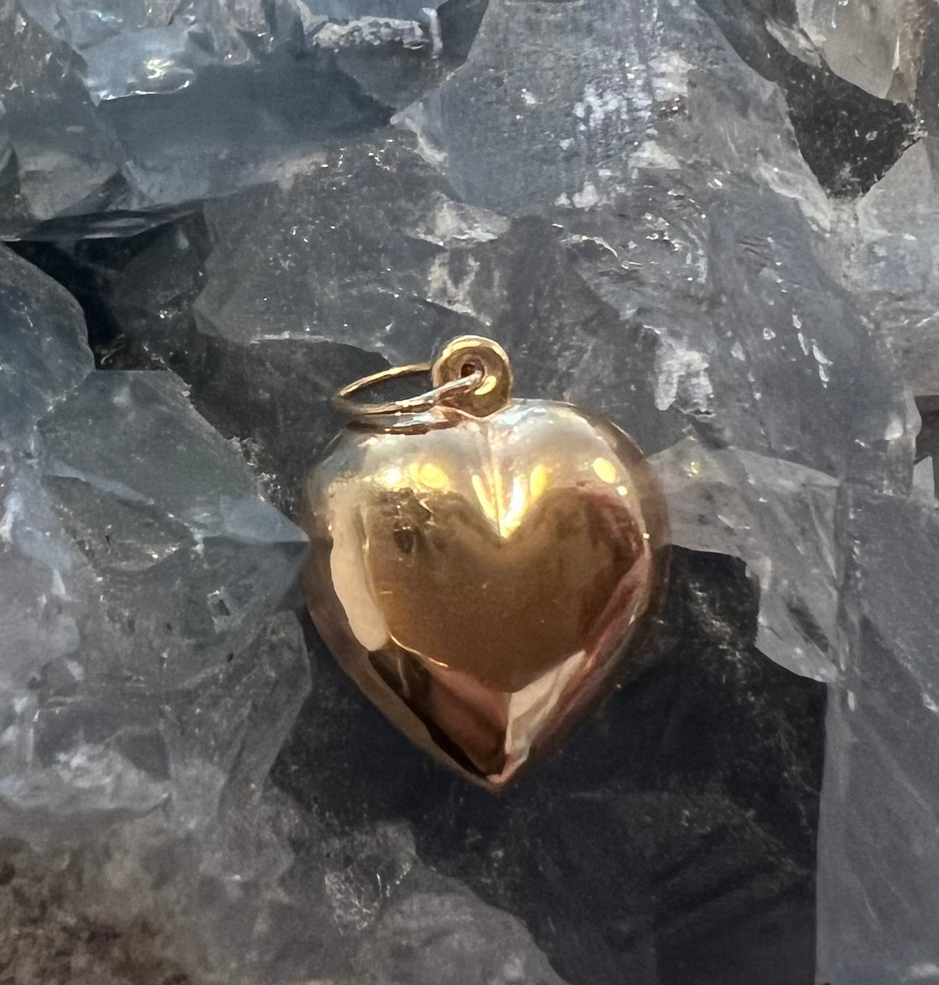 A polished large 14k gold organic heart-shaped pendant with a small loop at the top, resting on a bed of translucent gray crystals.