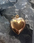 A polished large 14k gold organic heart-shaped pendant with a small loop at the top, resting on a bed of translucent gray crystals.