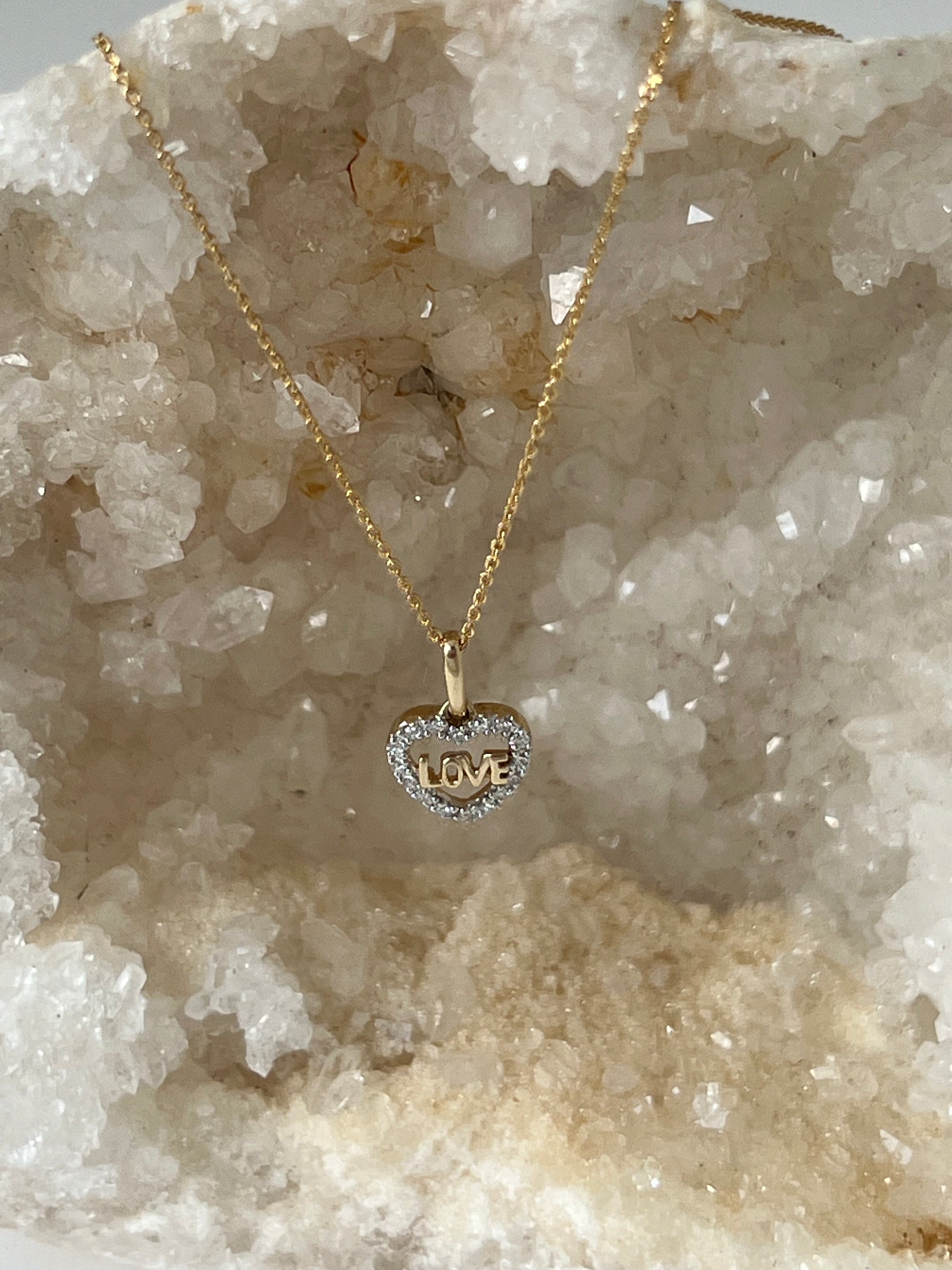 A solid 14k gold and diamond heart pendant necklace with the word 'LOVE' in the center, displayed on a crystal.